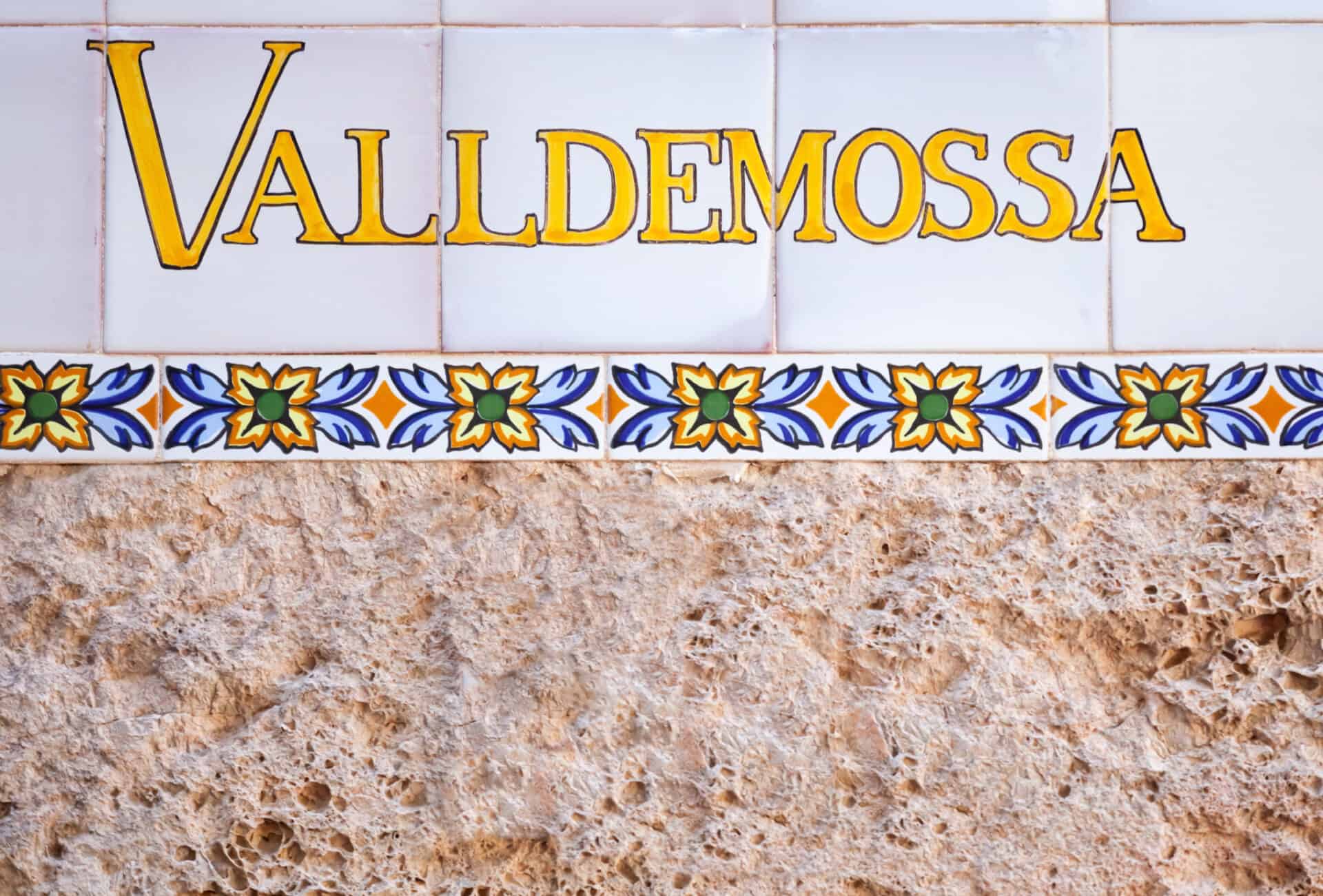 Ein handbemaltes spanisches Kachelschild für das Dorf Valldemossa (auf Katalanisch) oder Valldemosa (auf Spanisch), das in eine Steinmauer eingelassen ist.